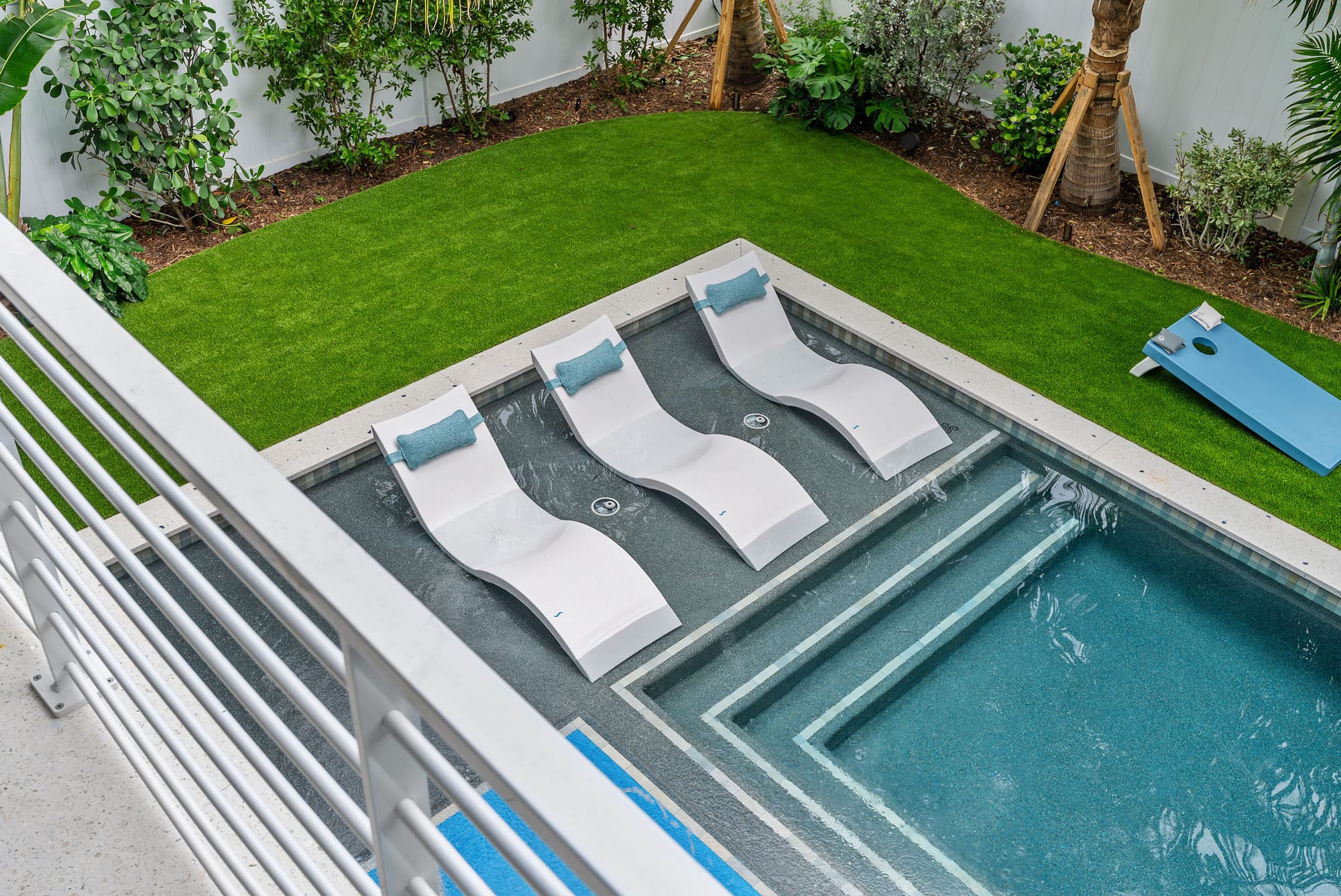 An overhead view of in-water loungers on a pool sun shelf bordered by lawn and tropical plantings.